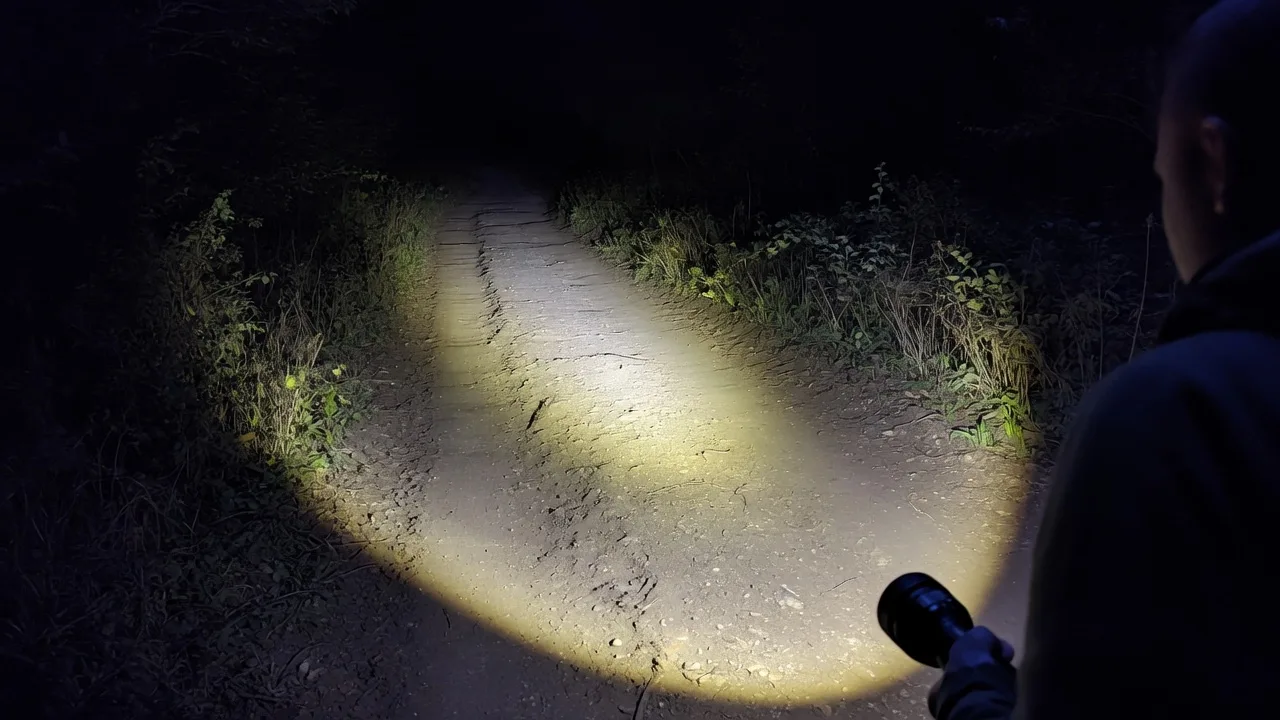 A person holding a flashlight that lights a rutted dirt trail with a wide pool of warm white light at night.